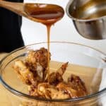 A hand pours sticky Mumbo Sauce recipe from a saucepan over crispy fried chicken pieces in a clear glass bowl, using a wooden spoon, on a wooden surface.