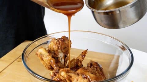 A hand pours sticky Mumbo Sauce recipe from a saucepan over crispy fried chicken pieces in a clear glass bowl, using a wooden spoon, on a wooden surface.