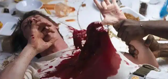 A man lies on a messy table covered in food, looking distressed as another person pours red liquid like blood onto his chest—a chaotic scene straight from a horror movie food moment and worthy of Bites of Terror.