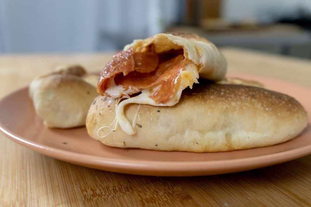 Two baked pepperoni rolls sit on a pink plate, one cut open to reveal gooey melted cheese and savory pepperoni slices inside, resting on a wooden surface with a blurred kitchen background.