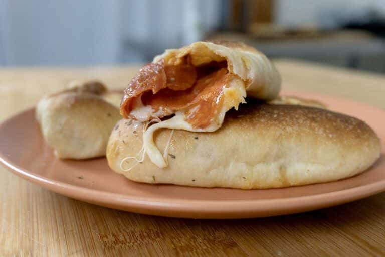 Two baked pepperoni rolls sit on a pink plate, one cut open to reveal gooey melted cheese and savory pepperoni slices inside, resting on a wooden surface with a blurred kitchen background.