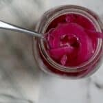 A spoon scoops vibrant Pickled Red Onions from a glass jar, viewed from above, on a white and gray marble surface with part of a striped cloth visible.