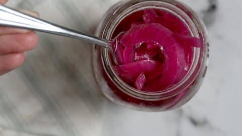 A spoon scoops vibrant Pickled Red Onions from a glass jar, viewed from above, on a white and gray marble surface with part of a striped cloth visible.