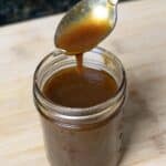 A glass jar filled with rich pumpkin spice syrup sits on a wooden surface; a spoon above the jar drips the fragrant sauce back inside.