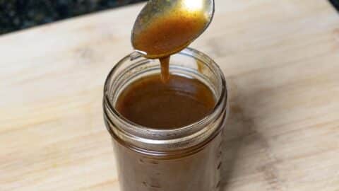 A glass jar filled with rich pumpkin spice syrup sits on a wooden surface; a spoon above the jar drips the fragrant sauce back inside.