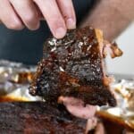 A person holds a glazed, cooked rib with shiny barbecue sauce, lifting it from a tray lined with aluminum foil and filled with more ribs.