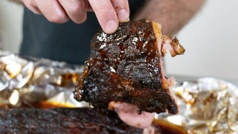 A person holds a glazed, cooked rib with shiny barbecue sauce, lifting it from a tray lined with aluminum foil and filled with more ribs.