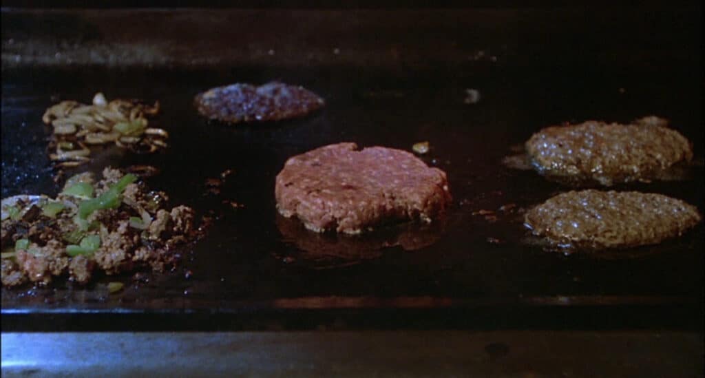 Raw and cooked hamburger patties, chopped onions, and green peppers are sizzling on a flat-top grill—almost like something out of one of those scary food scenes in Bites of Terror.