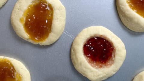 Close-up of Thumbprint Cookies on a baking sheet, filled with orange and red jam. The cookies have a light, crumbly texture and are arranged with visible space between them.