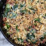 A cast iron skillet filled with a baked casserole inspired by the Utica Greens recipe, topped with golden, crispy breadcrumbs, melted cheese, leafy greens, and pieces of prosciutto or ham. The skillet rests on a white cloth.