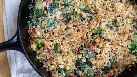 A cast iron skillet filled with a baked casserole inspired by the Utica Greens recipe, topped with golden, crispy breadcrumbs, melted cheese, leafy greens, and pieces of prosciutto or ham. The skillet rests on a white cloth.