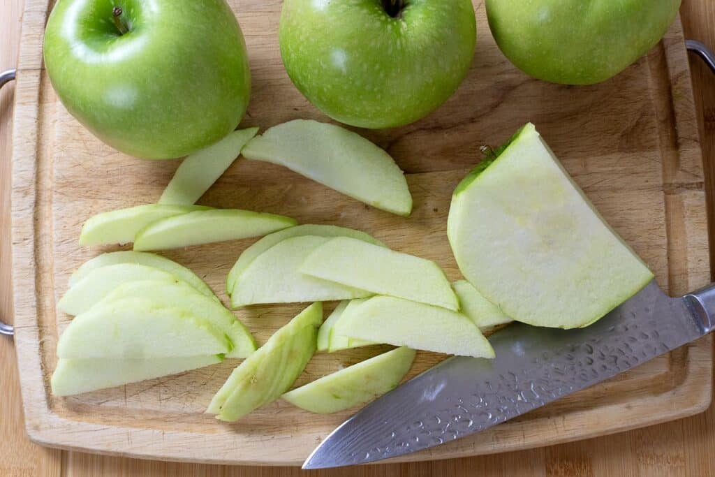 Three whole green apples and several thin apple slices are on a wooden cutting board. A large knife rests beside the slices. The setup is on a wooden surface.