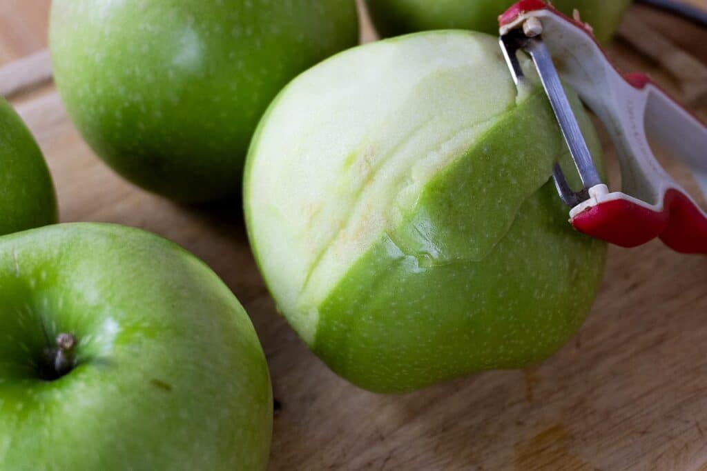 A close-up of a green apple being peeled with a red and white vegetable peeler on a wooden surface, with two more whole green apples nearby.