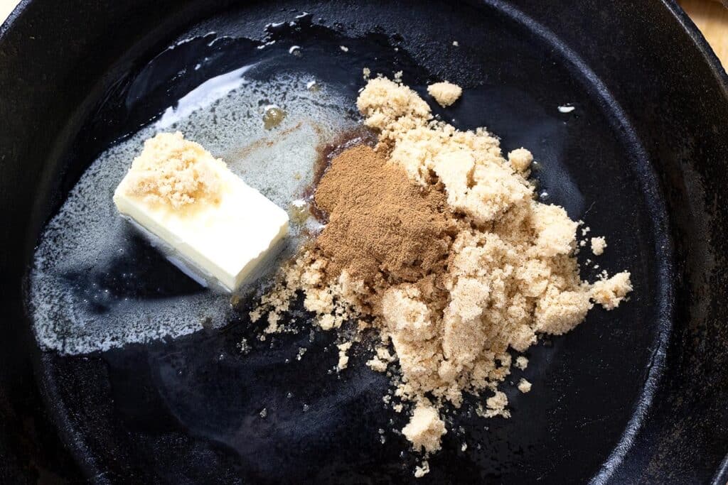 A cast iron skillet with a melting stick of butter, a pile of brown sugar, and a heap of cinnamon, all beginning to mix together.