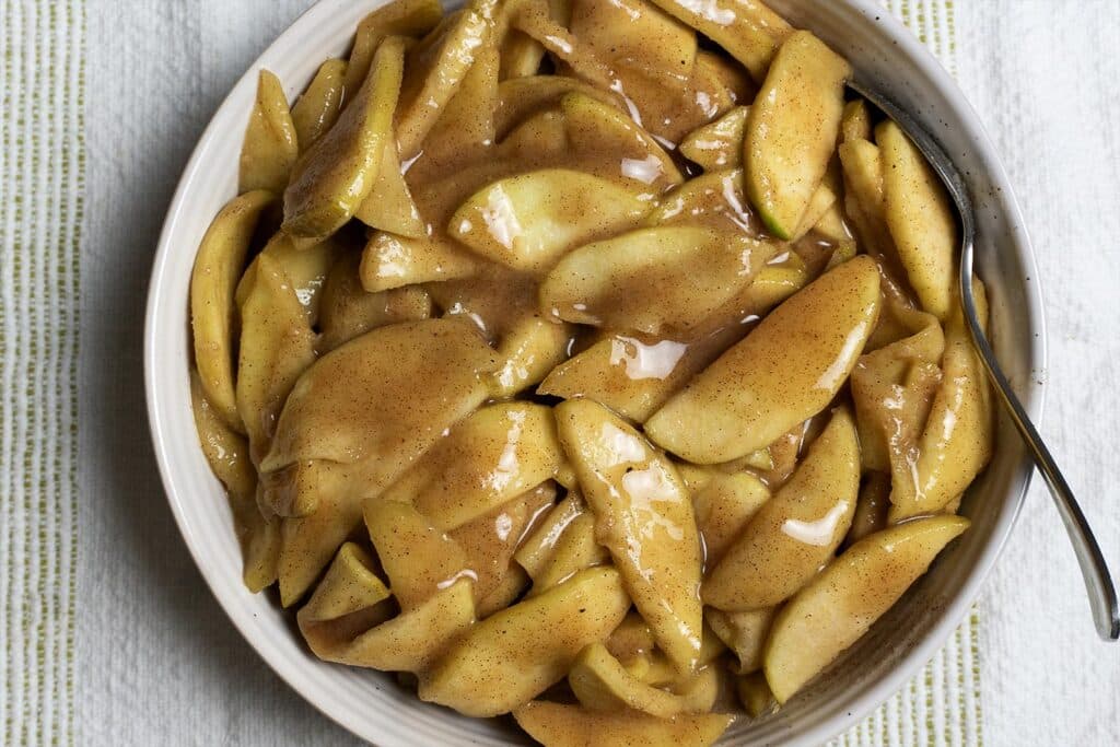 A bowl filled with sliced apples coated in a glossy cinnamon and sugar sauce, with a spoon resting on the side of the bowl, set on a striped cloth background.