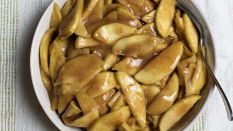 A bowl filled with sliced apples coated in a glossy cinnamon and sugar sauce, with a spoon resting on the side of the bowl, set on a striped cloth background.