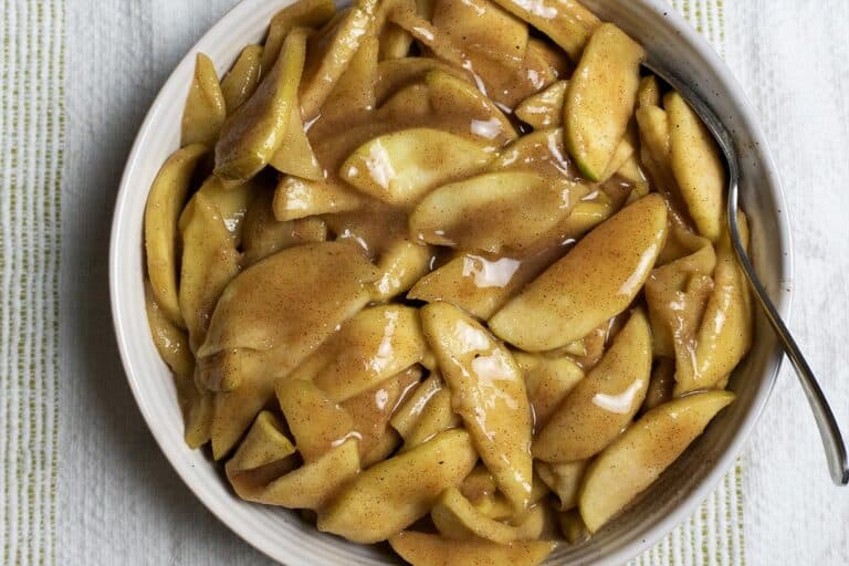 A bowl filled with sliced apples coated in a glossy cinnamon and sugar sauce, with a spoon resting on the side of the bowl, set on a striped cloth background.