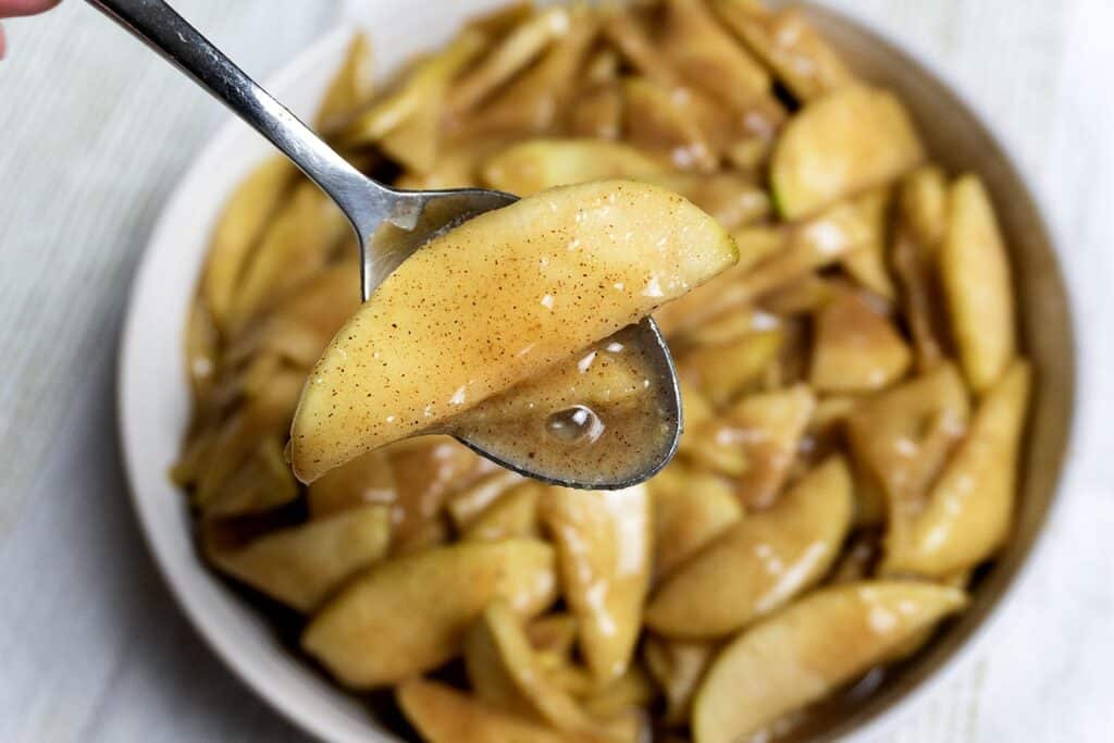 A close-up of a spoon holding glazed apple slices with cinnamon, above a bowl filled with more of the same apple slices in syrup.