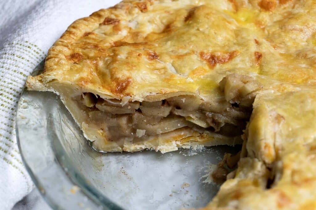 A close-up of a homemade apple pie in a glass pie dish, with a slice missing. The golden-brown crust covers layers of sliced apples inside. A white kitchen towel with green stripes is visible in the background.