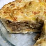 A close-up of a homemade apple pie in a glass pie dish, with a slice missing. The golden-brown crust covers layers of sliced apples inside. A white kitchen towel with green stripes is visible in the background.
