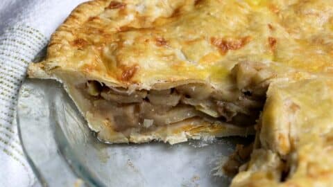 A close-up of a homemade apple pie in a glass pie dish, with a slice missing. The golden-brown crust covers layers of sliced apples inside. A white kitchen towel with green stripes is visible in the background.