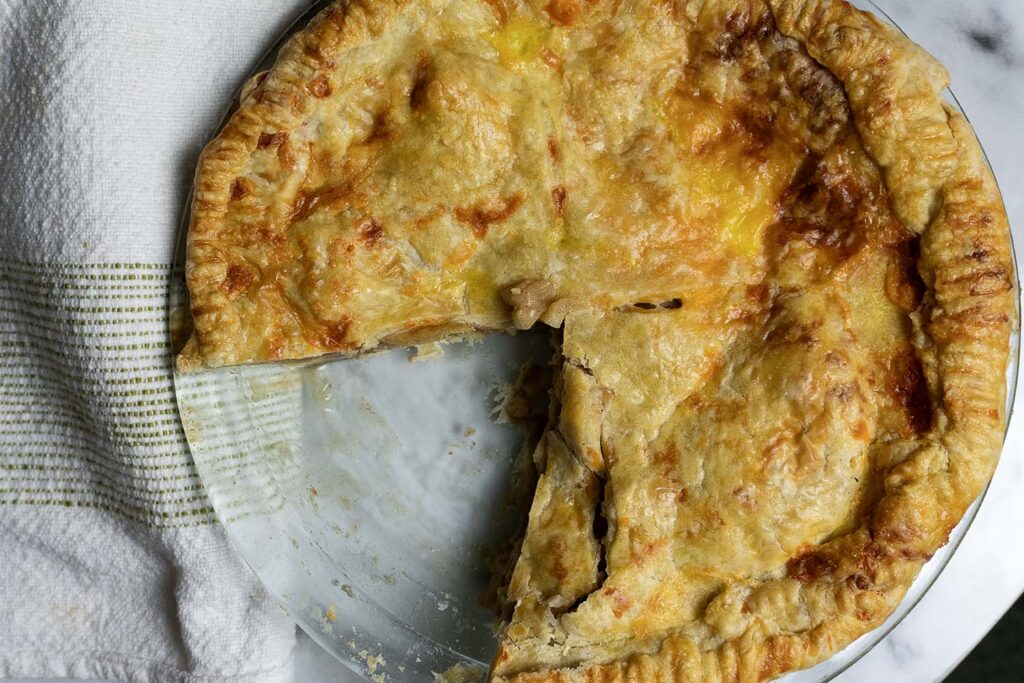 A golden-brown pie with a flaky crust sits in a glass dish. One slice has been removed, revealing the filling inside. A white towel with green stripes is partially visible beside the pie.