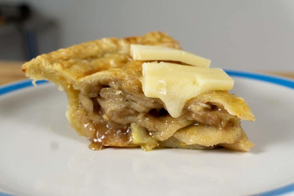 A slice of apple pie with a flaky crust, topped with two slices of butter, served on a white plate with a blue rim.