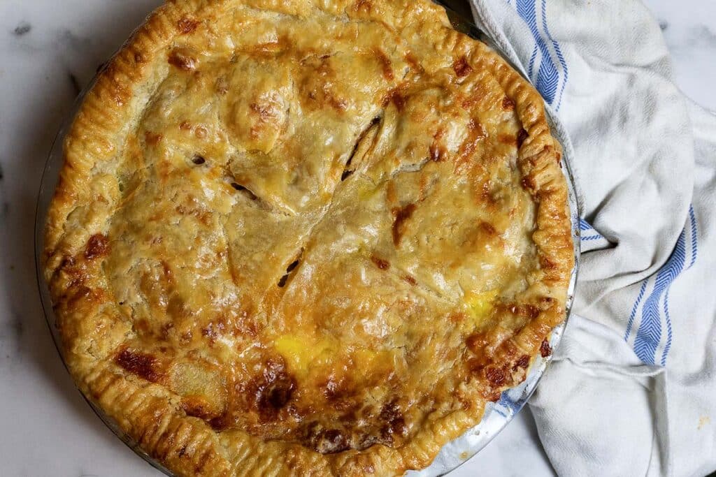 A golden-brown, freshly baked pie with a flaky crust sits on a marble surface next to a white cloth with blue stripes.