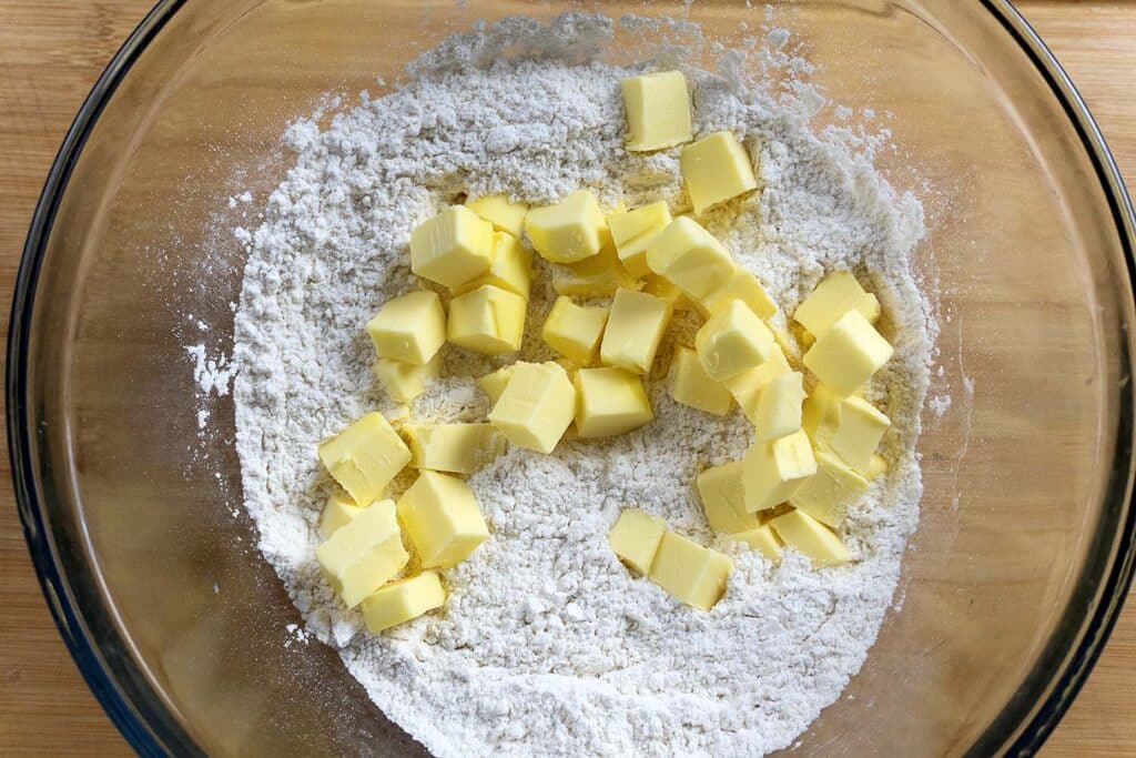A glass bowl containing flour with small cubes of butter on top, ready to be mixed on a wooden surface.