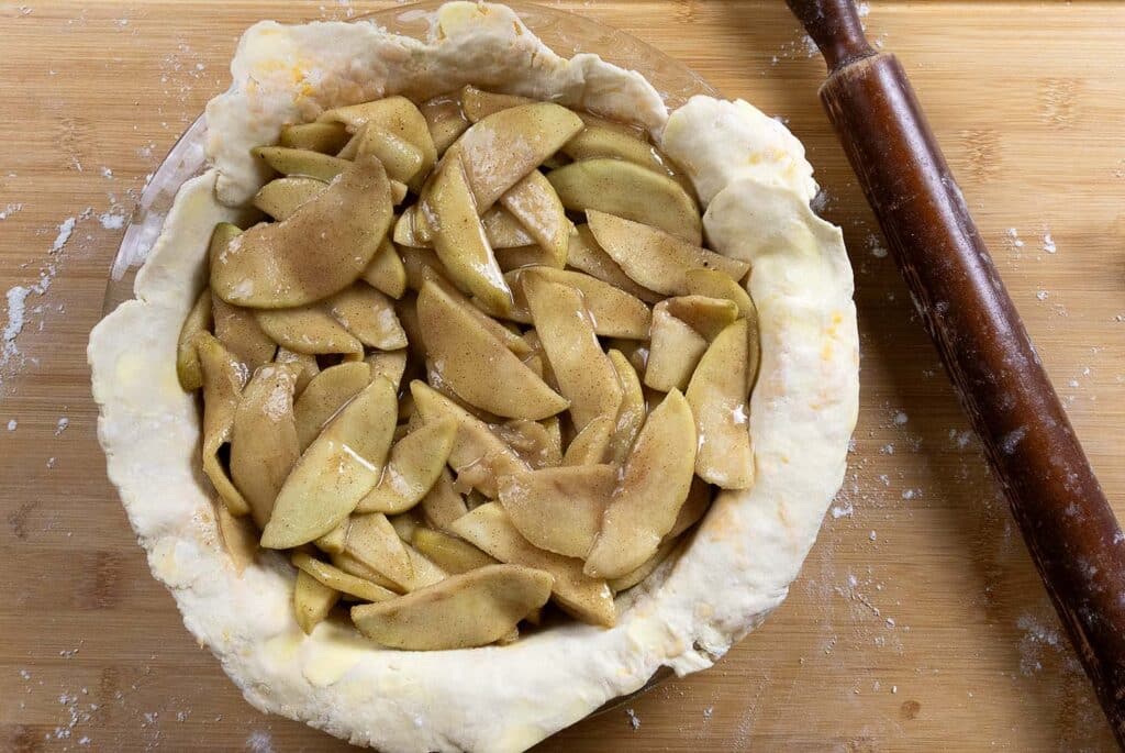 A pie dish filled with sliced apples in cinnamon and sugar sits on an unbaked pie crust, with a rolling pin nearby on a wooden surface dusted with flour.