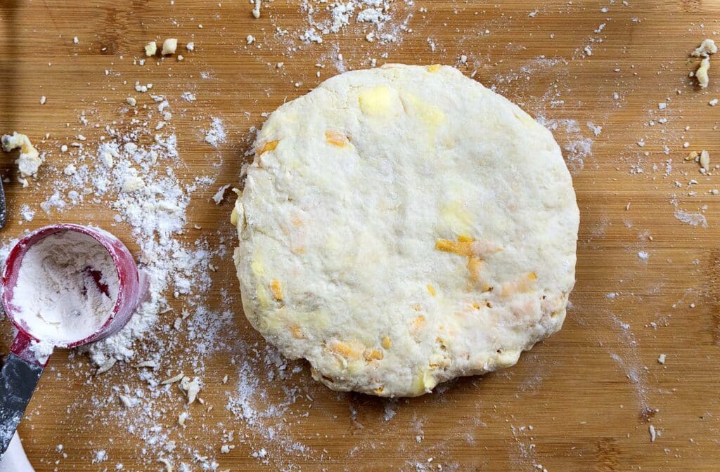 A round, flattened ball of biscuit dough with visible bits of butter sits on a floured wooden surface next to a red measuring cup with flour. Scattered flour and dough bits are around.