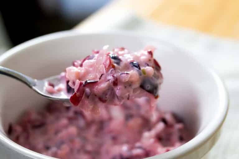 A close-up of a spoonful of creamy, pinkish cranberry sauce with chunks of fruit being lifted from a white bowl. The salad has a mixed texture with visible pieces of red and purple fruit.