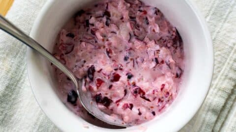 A bowl of creamy, pinkish cranberry sauce mixed with visible chunks of cranberries, with a spoon resting inside. The bowl is placed on a textured, off-white cloth.