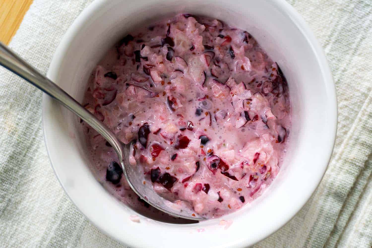 A bowl of creamy, pinkish cranberry sauce mixed with visible chunks of cranberries, with a spoon resting inside. The bowl is placed on a textured, off-white cloth.