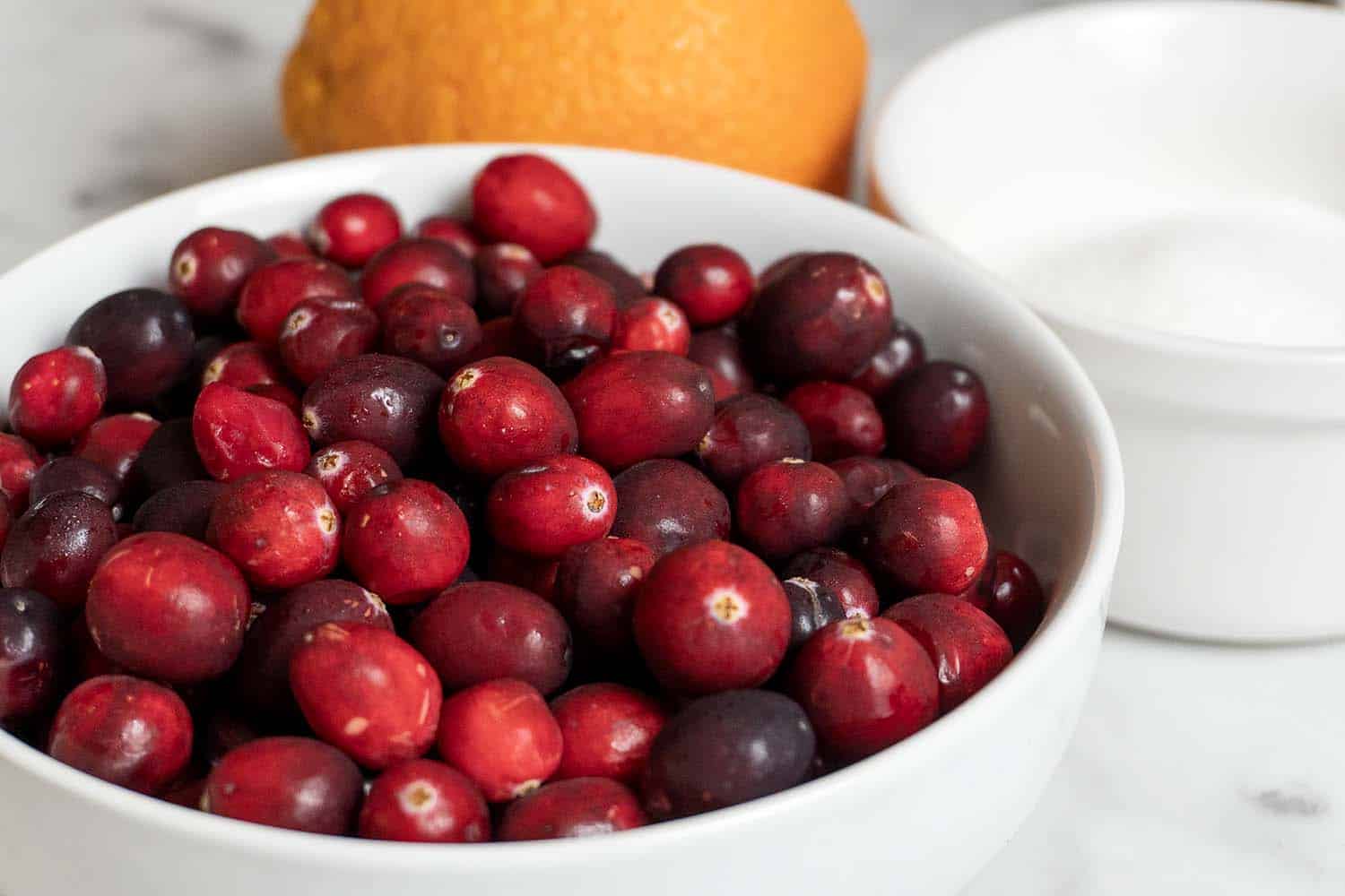 A white bowl filled with fresh cranberries sits on a marble surface, with a whole orange and a small bowl of sugar in the background.