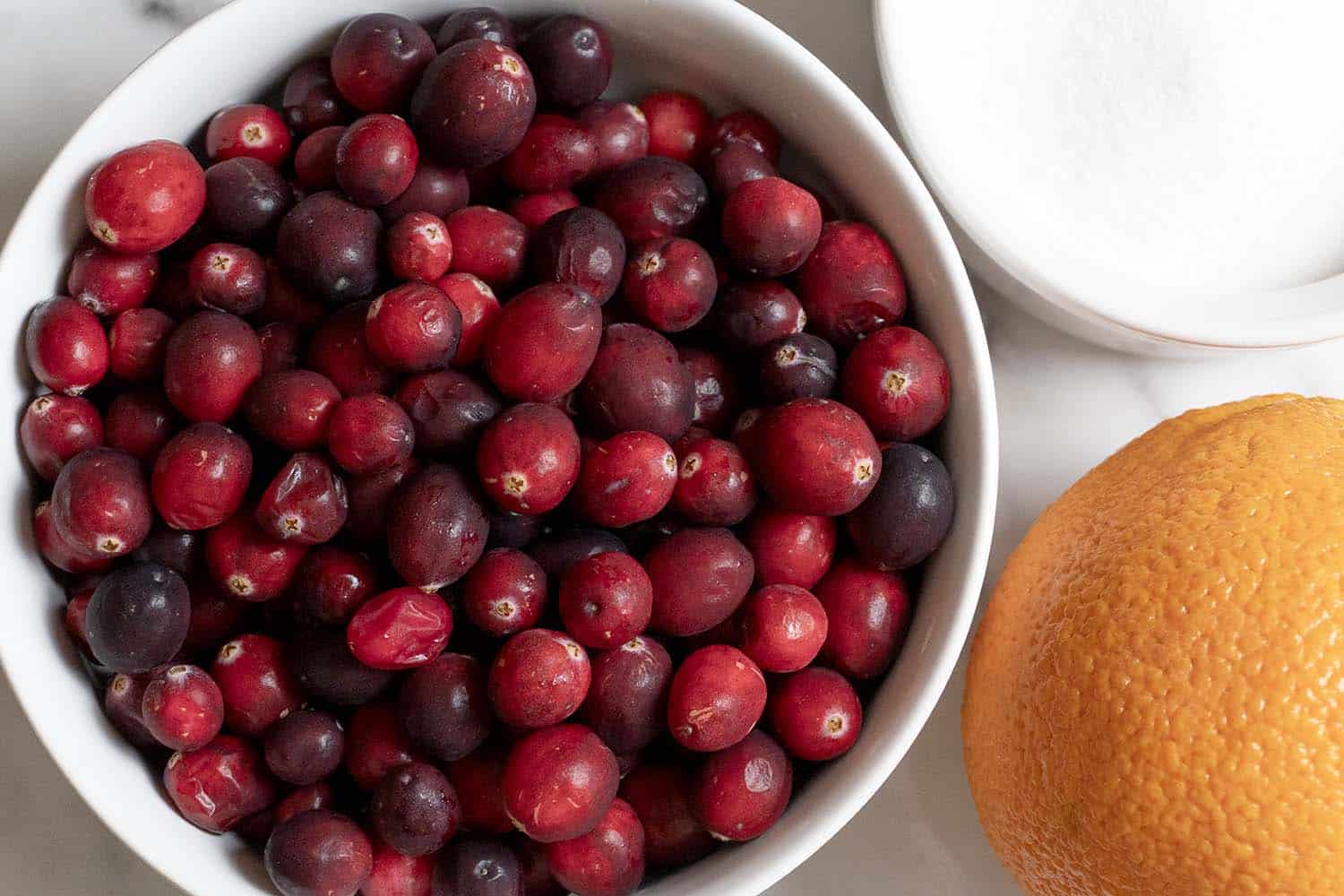 A bowl of fresh cranberries next to a whole orange and a bowl of white sugar, all placed on a white surface.