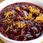A white bowl filled with homemade cranberry sauce, topped with freshly grated orange zest, sits on a wooden surface. The sauce is deep red and chunky in texture.