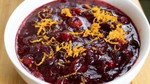 A white bowl filled with homemade cranberry sauce, topped with freshly grated orange zest, sits on a wooden surface. The sauce is deep red and chunky in texture.