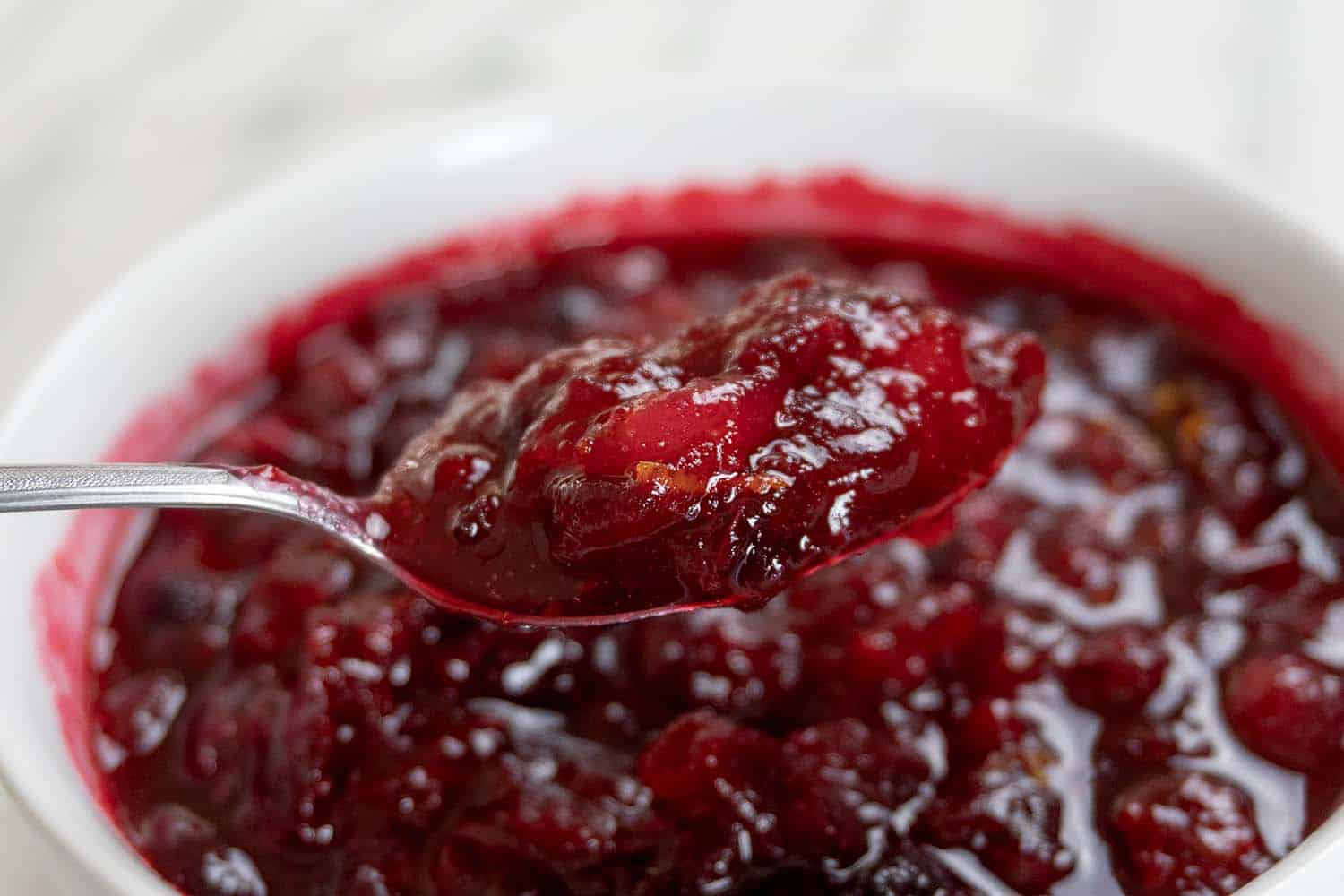 A close-up of a spoonful of chunky, red cranberry sauce being lifted from a white bowl filled with more cranberry sauce.