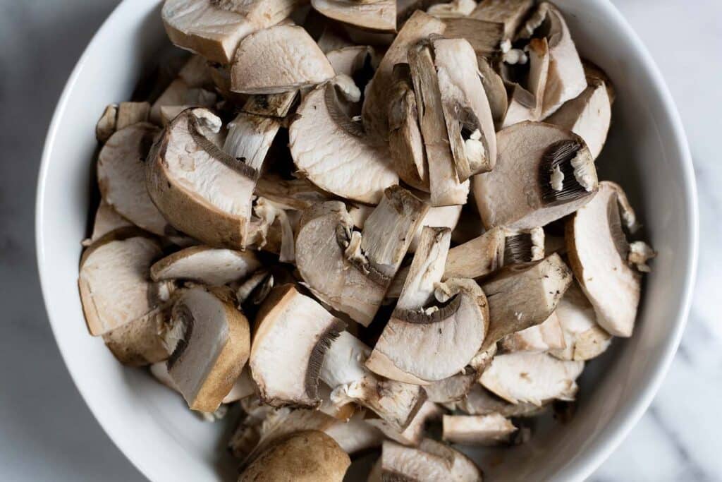 A white bowl filled with chopped brown mushrooms, showing their caps and stems, placed on a white surface.