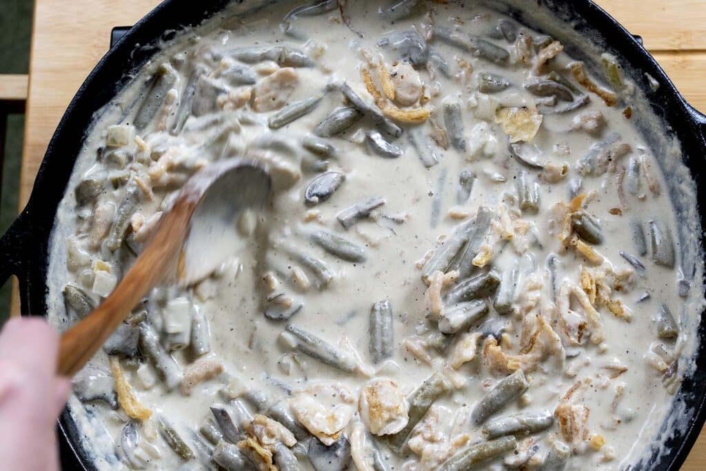 A close-up of a person stirring creamy green bean casserole in a black skillet with a wooden spoon. The casserole contains green beans, onions, and a thick white sauce.