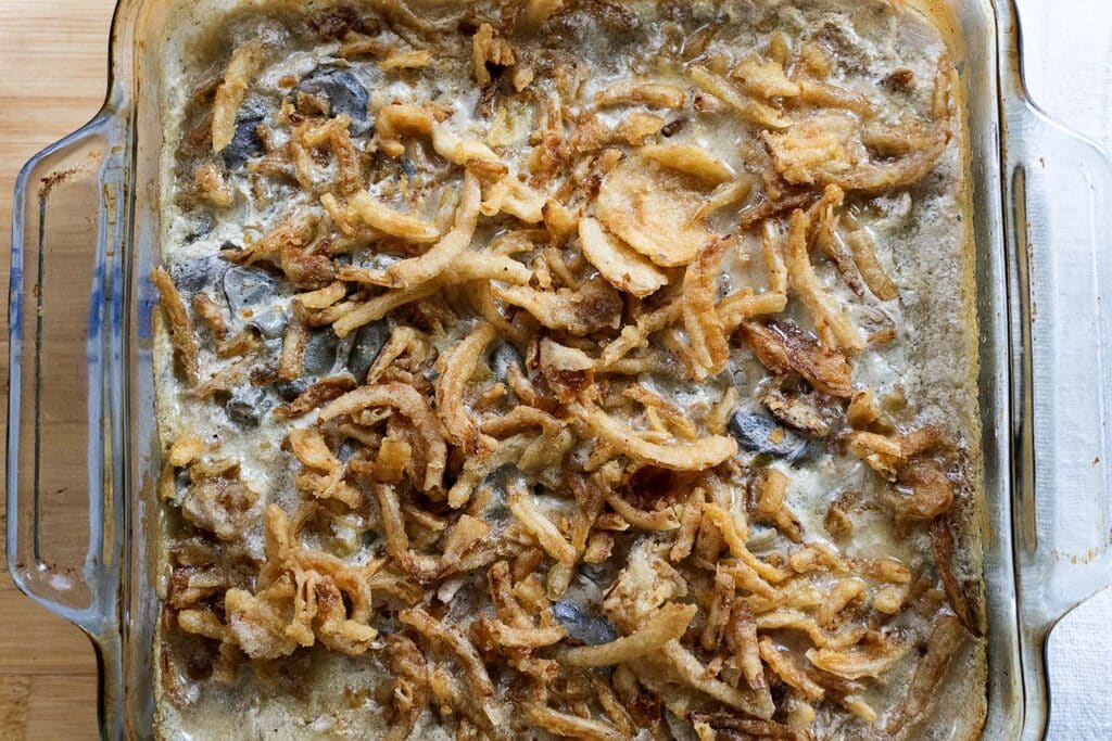 A glass baking dish filled with green bean casserole, topped with crispy fried onions and a creamy sauce, resting on a wooden surface.