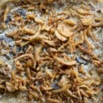 A glass baking dish filled with green bean casserole, topped with crispy fried onions and a creamy sauce, resting on a wooden surface.