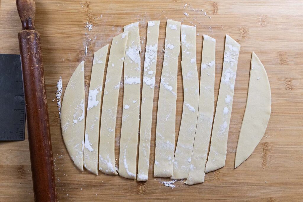 Thin strips of dough dusted with flour are laid out on a wooden surface, beside a rolling pin and a metal scraper. The dough appears to be freshly rolled and cut into even pieces.