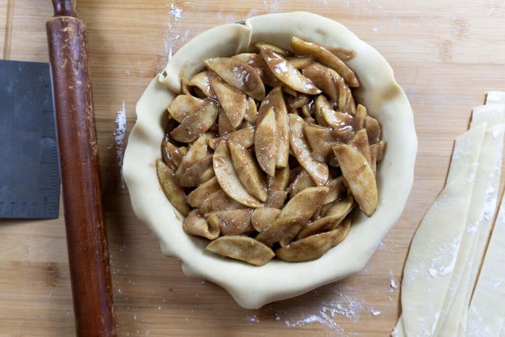 A pie crust filled with spiced apple slices sits on a wooden surface, ready to be covered. A rolling pin and strips of dough are nearby, along with some scattered flour.