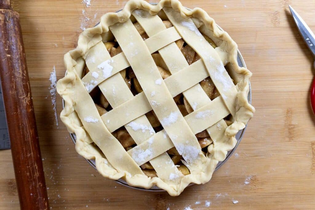Unbaked apple pie with a lattice crust, sprinkled with flour, sits on a wooden surface next to a rolling pin and a pair of scissors.