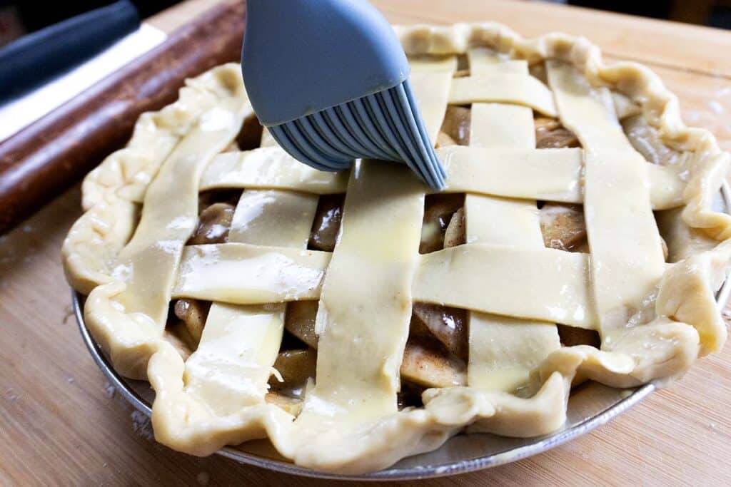 A close-up of an unbaked lattice-topped pie being brushed with an egg wash using a silicone pastry brush, set on a wooden surface with a rolling pin in the background.