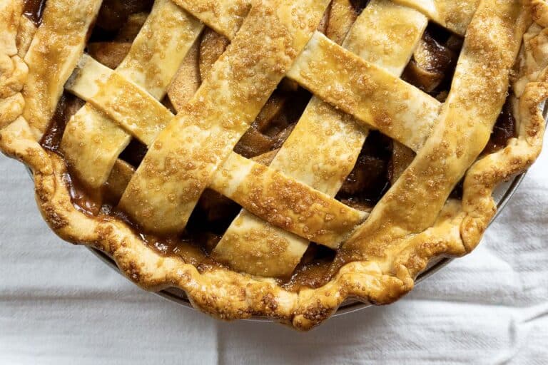 A close-up of a golden-brown lattice-topped pie with a flaky crust and visible sugar crystals, sitting on a white cloth. The pie filling appears to be fruit, likely apple, with caramelized edges.