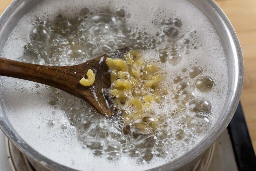 A wooden spoon stirs macaroni pasta boiling in a pot of water, with bubbles and foam rising to the surface on a stovetop.