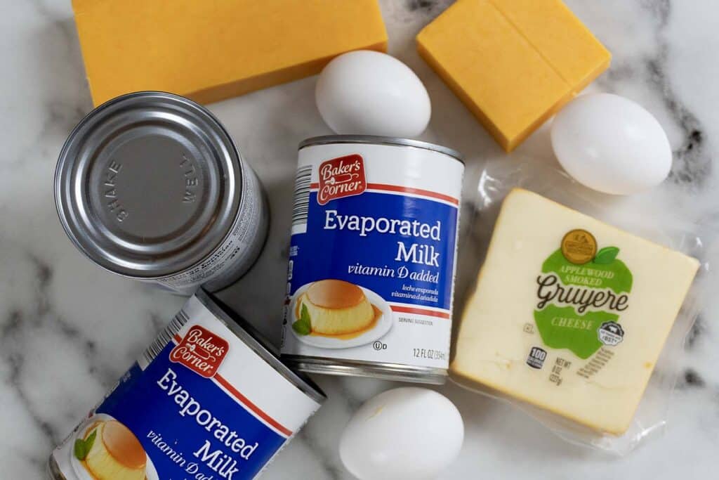 Two cans of evaporated milk, three eggs, two blocks of orange cheese, and a package of sliced Gruyere cheese are arranged on a white marble surface.